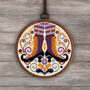 May include: A circular embroidery hoop with a white background and a colorful design of two witch's boots with a purple, orange, and black color scheme. The design is surrounded by a decorative pattern of swirls and leaves in shades of gray, orange, and purple.