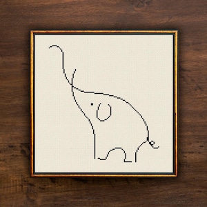 May include: A framed cross-stitch artwork featuring a minimalist black line drawing of an elephant. The elephant is depicted with a raised trunk and a small, circular eye. The artwork is set against a cream-colored background and framed in gold.