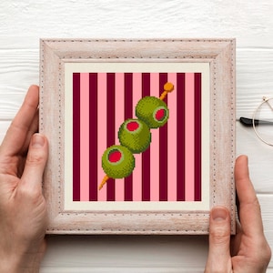 May include: A square framed cross-stitch embroidery featuring a pixel art design of three green olives with red centers on a yellow toothpick, set against a background of alternating vertical stripes in shades of light pink and dark maroon.