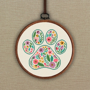 Może przedstawiać: Obręcz do haftu krzyżykowego z białym tłem z tkaniny przedstawiającą kwiatowy wzór odcisku łapy. Odcisk łapy jest obwiedziony turkusowym kolorem i wypełniony różowymi, fioletowymi, żółtymi i zielonymi kwiatami.