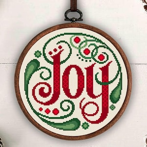 May include: A circular wooden embroidery hoop containing a cross-stitch design of the word Joy in red, surrounded by decorative green swirls and small red dots. The hoop is suspended by a brown ribbon at the top, centered against a white background.