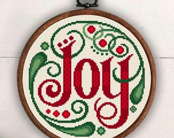 May include: A circular wooden embroidery hoop containing a cross-stitch design of the word Joy in red, surrounded by decorative green swirls and small red dots. The hoop is suspended by a brown ribbon at the top, centered against a white background.