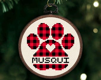 May include: A round wooden embroidery hoop ornament featuring a cross-stitched red and black buffalo plaid paw print design. The center of the paw contains a small heart cutout, and the name MUSQUI is stitched in black letters at the bottom.
