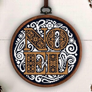 May include: A circular wooden embroidery hoop containing a cross-stitch pattern on a dark blue fabric. The design features the word NOEL in large, tan, patterned block letters surrounded by intricate white floral and swirling scrollwork.
