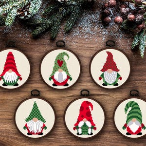 May include: Six cross-stitch Christmas ornaments with gnome designs. Each ornament has a wooden hoop and a small metal loop for hanging. The gnomes have red, green, and white hats and beards, with green and red outfits.