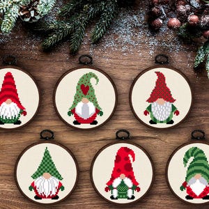 May include: Six circular embroidery hoops containing cross-stitch patterns of gnomes. Each gnome features a unique hat design in red and green, a white beard, and a colorful outfit. The hoops are framed in dark wood and have black hanging loops.