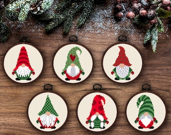 May include: Six circular embroidery hoops containing cross-stitch patterns of gnomes. Each gnome features a unique hat design in red and green, a white beard, and a colorful outfit. The hoops are framed in dark wood and have black hanging loops.