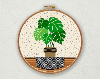 May include: A cross-stitch pattern of a potted green monstera plant on a black and white patterned rug. The plant is in a brown pot with a wooden base. The background is a beige fabric with a speckled pattern.