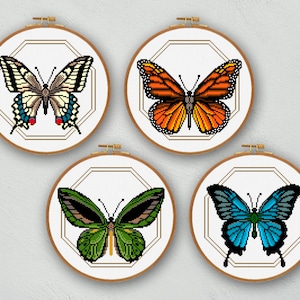 May include: Four framed cross-stitch butterfly designs. Each butterfly is displayed in a round wooden hoop with a geometric gold border. The butterflies feature various colours, including orange, blue, green, and black.