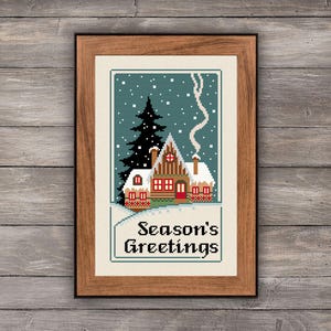 May include: A framed cross-stitch embroidery featuring a winter scene with a snow-covered house, a tall black pine tree, and falling white snowflakes against a teal background. The text Season's Greetings is stitched in black at the bottom.