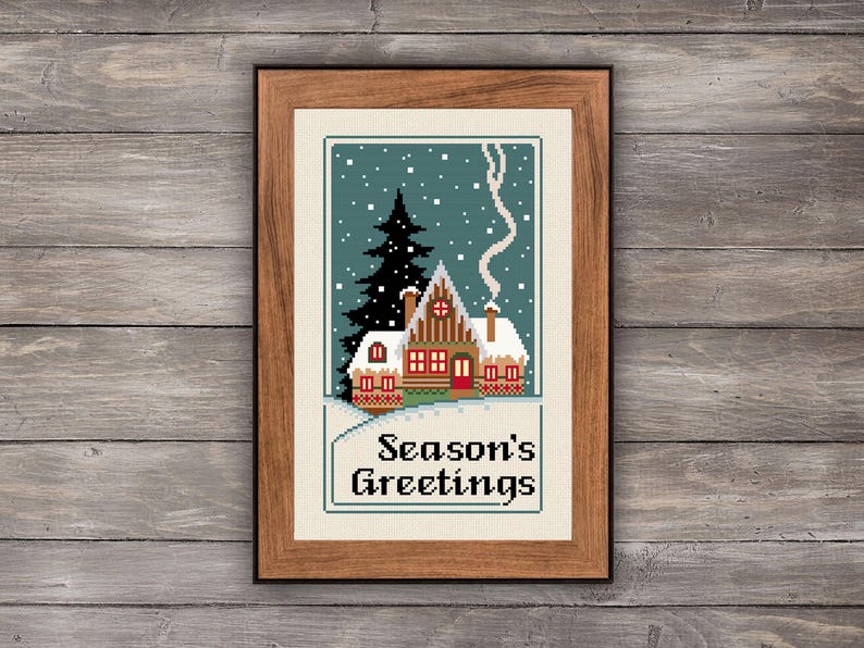 May include: A framed cross-stitch embroidery featuring a winter scene with a snow-covered house, a tall black pine tree, and falling white snowflakes against a teal background. The text Season's Greetings is stitched in black at the bottom.