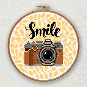 Può includere: Uno schema di punto croce con una macchina fotografica vintage di colore marrone e grigio, circondata da foglie gialle e la parola "Smile".