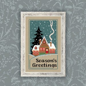 May include: A framed cross-stitch embroidery featuring a winter cabin scene with a tall dark green pine tree, snow-covered roof, and smoke rising from a chimney. The text "Season's Greetings" is stitched in black at the bottom on a tan fabric background.