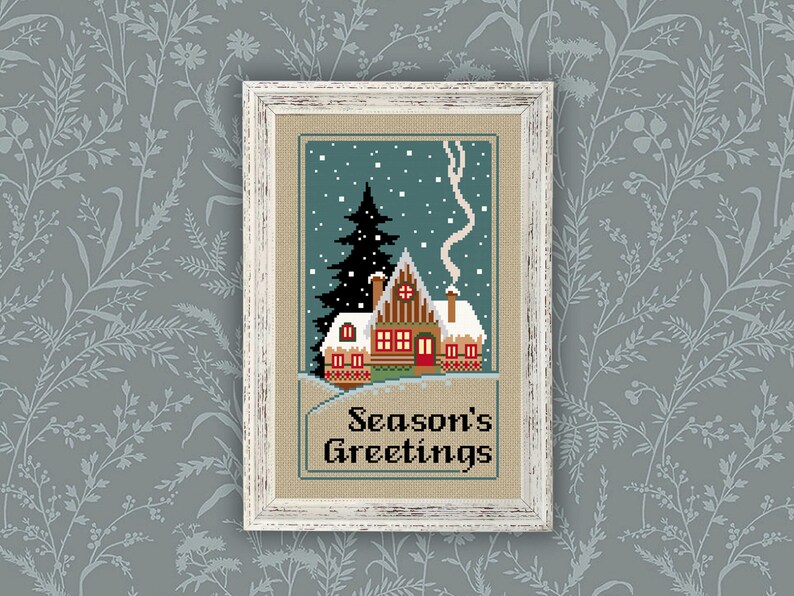 May include: A framed cross-stitch embroidery featuring a winter cabin scene with a tall dark green pine tree, snow-covered roof, and smoke rising from a chimney. The text "Season's Greetings" is stitched in black at the bottom on a tan fabric background.