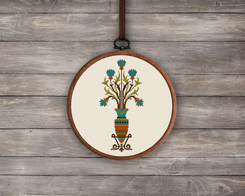 May include: A cross-stitch embroidery hoop with a design of a vase with blue flowers and green leaves. The vase is decorated with brown, orange, and blue stripes.