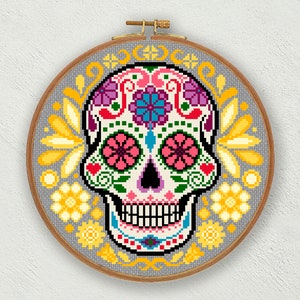 May include: A cross-stitch sugar skull design in a wooden hoop. The skull is decorated with floral patterns in pink, purple, blue, and yellow, set against a grey background. The embroidery hoop is approximately 20 cm in diameter.