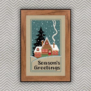 May include: A framed cross-stitch embroidery featuring a winter scene with a snow-covered cabin, a tall dark pine tree, and falling snow against a teal sky. The text Season's Greetings is stitched in black at the bottom of the tan fabric.