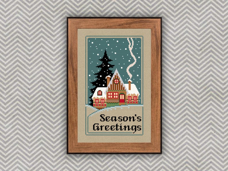May include: A framed cross-stitch embroidery featuring a winter scene with a snow-covered cabin, a tall dark pine tree, and falling snow against a teal sky. The text Season's Greetings is stitched in black at the bottom of the tan fabric.