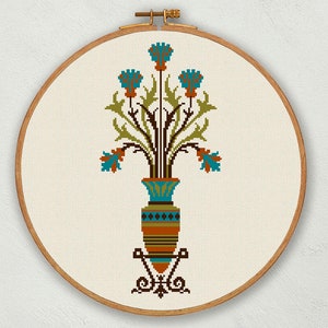May include: A cross-stitch embroidery design of a vase with blue flowers and green leaves. The vase is brown and orange with a blue and white pattern. The design is framed in a wooden hoop.