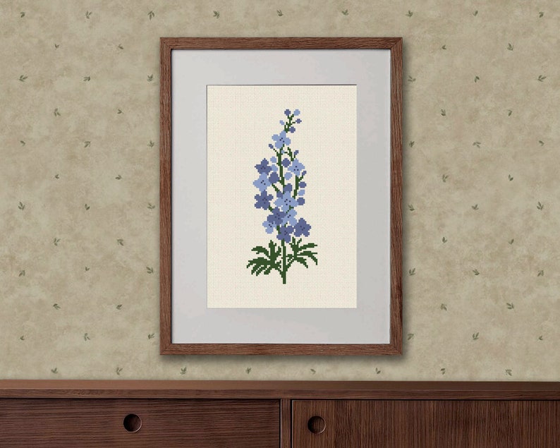 以下が含まれることがあります： 青い花と緑の葉のデルフィニウムの花のクロスステッチ刺繍が額装されています