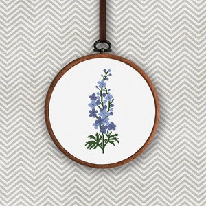 以下が含まれることがあります： 青い花柄のクロスステッチ刺繍枠。花は薄い青色で、緑色の茎と葉があります。