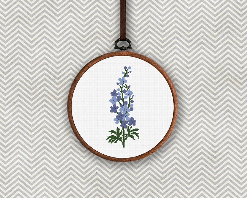 以下が含まれることがあります： 青い花柄のクロスステッチ刺繍枠。花は薄い青色で、緑色の茎と葉があります。