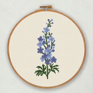 以下が含まれることがあります： 木製の刺繍枠に入れた、青と緑のデルフィニウムの花のクロスステッチパターンです。