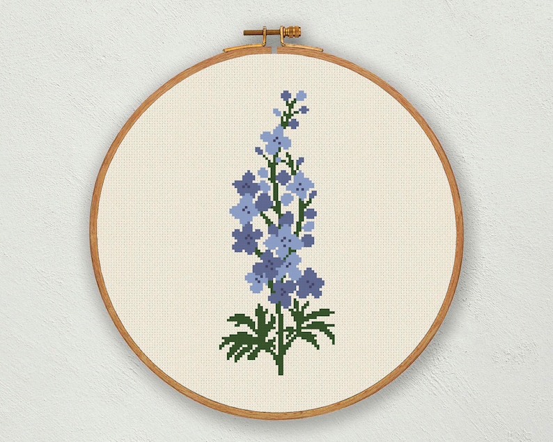 以下が含まれることがあります： 木製の刺繍枠に入れた、青と緑のデルフィニウムの花のクロスステッチパターンです。