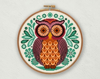 May include: A circular wooden embroidery hoop containing a cross-stitch pattern of a stylized owl. The owl features a dark red face, orange eyes, and a patterned orange chest. It is surrounded by teal floral and leaf motifs on a white fabric.