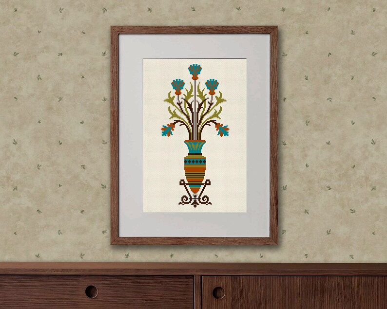 May include: A framed cross-stitch embroidery of a stylized vase with blue flowers and green leaves. The vase is brown, orange, and blue with a geometric pattern.