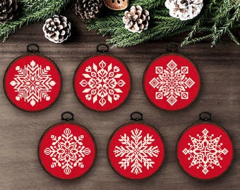 May include: Six circular embroidery hoops featuring red fabric with white cross-stitched snowflake patterns. Each hoop has a dark brown wooden frame and a small black metal hanging loop at the top, displaying unique geometric snowflake designs.