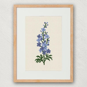 以下が含まれることがあります： 青い花と緑の葉のデルフィニウムの花のクロスステッチ刺繍が額装されています