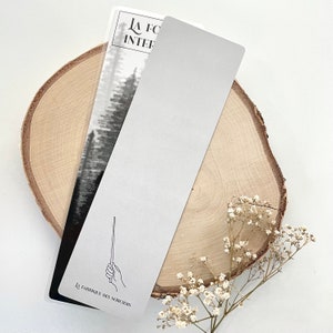 May include: Two rectangular bookmarks. One features a black and white forest scene, the other is plain silver. The first bookmark has text that reads "LA FOR&Ecirc;T INTERIEURE" and "LA FABRIQUE DES SORCIERS" with a wand illustration. The bookmarks rest on a wooden slice.
