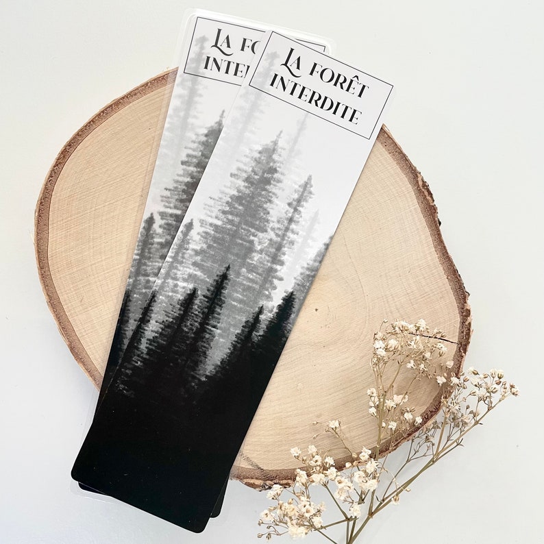 May include: Two bookmarks with a black and white forest design. The bookmarks have the text "LA FOR&Ecirc;T INTERDITE" at the top. The bookmarks are on a wooden slice with small white flowers.
