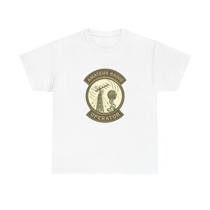 Puede incluir: Camiseta blanca con un diseño circular. El diseño incluye las palabras "AMATEUR RADIO OPERATOR" y presenta una torre de radio, una antena y un micrófono en tonos marrones y beige.