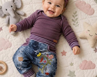 Puede incluir: Un bebé viste una camiseta de manga larga morada con puños y cintura de rayas blancas, combinada con pantalones vaqueros azules de lavado ácido con un bordado colorido de una planta y un pájaro. Lleva una diadema morada a juego con un lazo.