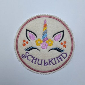 Könnte beinhalten: Ein runder, cremefarbener Filz-Patch mit einem aufgestickten pinkfarbenen Einhorn-Gesicht. Das Einhorn hat ein Regenbogenhorn, rosa Ohren und schwarze Wimpern. Der Text "SCHULKIND" ist in Lila unterhalb des Einhorns gestickt.