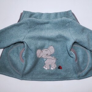 Könnte beinhalten: Eine tealblaue Fleecejacke mit Reißverschluss und zwei Taschen. Die Jacke hat ein gesticktes Elefantenmotiv auf der Rückseite mit einem Marienkäfer.