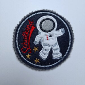 Könnte beinhalten: Ein runder, grauer Filz-Patch mit einem weißen Astronauten in einem Raumanzug, rotem Text, der "Schulkend" sagt, und drei goldenen Sternen.
