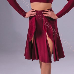 May include: A woman wearing a burgundy dance outfit with a long-sleeved crop top and a skirt with a slit and fringe detail. She is wearing brown dance shoes.