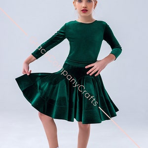 Pode incluir: Uma pessoa jovem vestindo um vestido de dança de veludo verde com mangas compridas e saia rodada. Ela está usando meias brancas e sapatos brancos com uma tira.