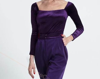 Puede incluir: Una mujer con un top de terciopelo morado de manga larga y pantalones anchos a juego. Está de pie con la mano en el bolsillo y mirando a la cámara.