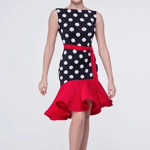 Op de afbeelding: Een zwart-witte polkadotjurk met een rode geruite zoom en een rode sash. De jurk is mouwloos en heeft een ronde hals.