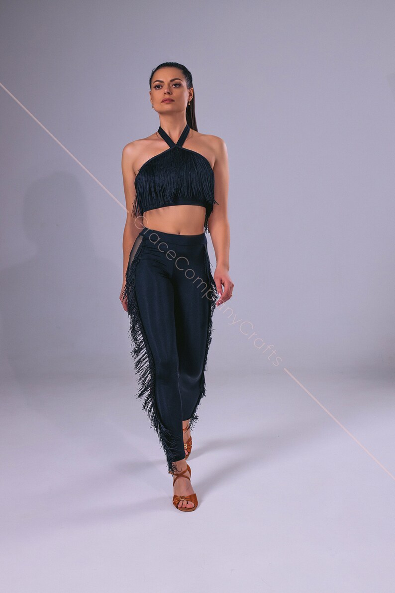 May include: A black halter top and matching pants with fringe trim. The top is a cropped style and the pants are fitted. The model is wearing brown sandals.