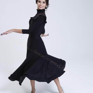 May include: A woman wearing a black mock neck dress with a ruffled neckline and a flowing skirt. She is standing on a white background and is wearing silver shoes.