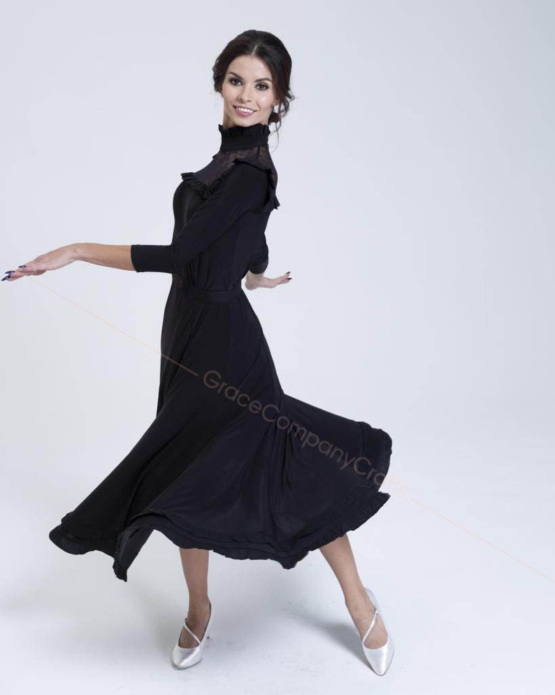 May include: A woman wearing a black mock neck dress with a ruffled neckline and a flowing skirt. She is standing on a white background and is wearing silver shoes.