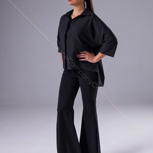 Puede incluir: Una mujer con una camisa negra de gran tama&ntilde;o con cuello y mangas largas, pantalones negros acampanados y sandalias marrones. La camisa est&aacute; metida en los pantalones.