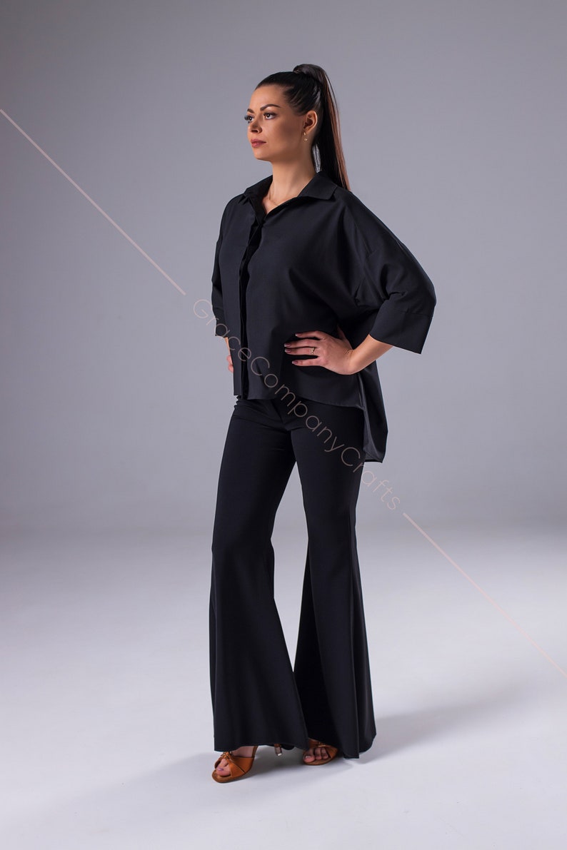 Puede incluir: Una mujer con una camisa negra de gran tama&ntilde;o con cuello y mangas largas, pantalones negros acampanados y sandalias marrones. La camisa est&aacute; metida en los pantalones.
