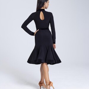 K&ouml;nnte beinhalten: Ein schwarzes Tanzkleid mit Stehkragen, langen &Auml;rmeln und einem figurbetonten Oberteil. Das Kleid hat einen ausgestellten Rock mit einem Samtbesatz. Das Model tr&auml;gt braune Schuhe mit Absatz.
