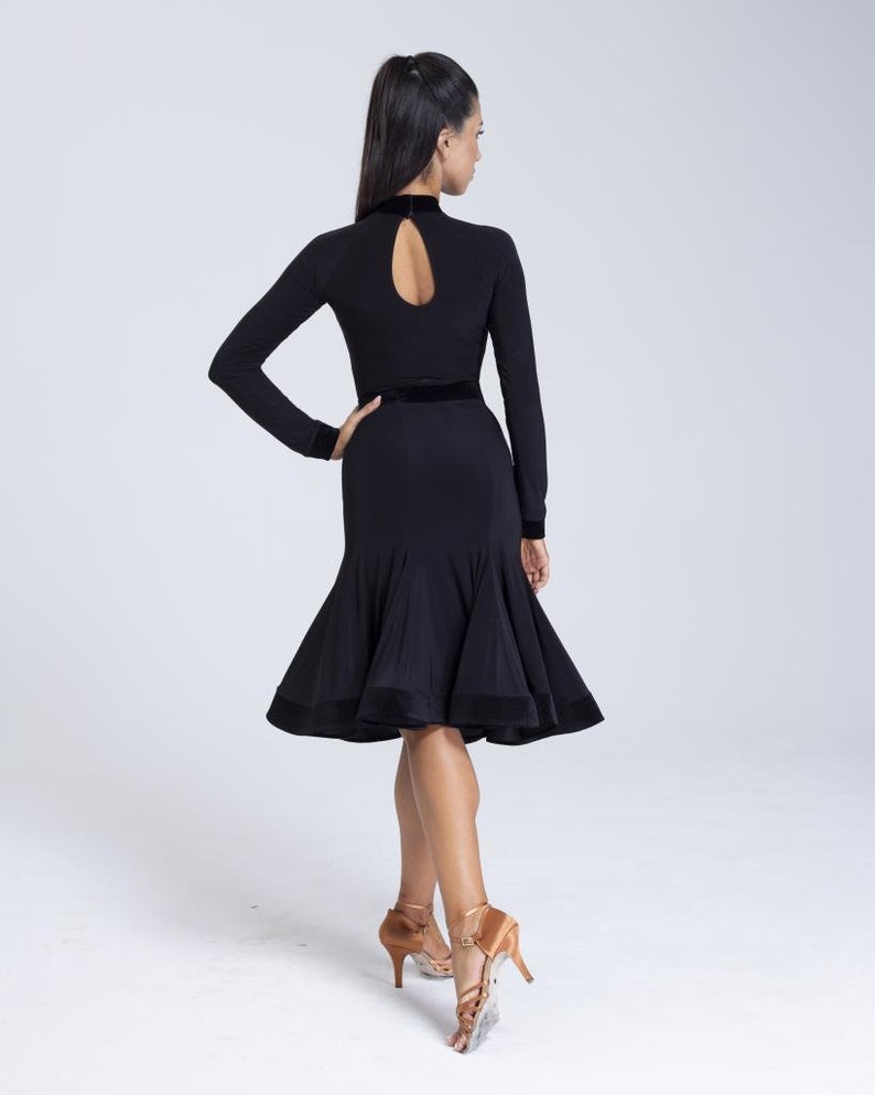 K&ouml;nnte beinhalten: Ein schwarzes Tanzkleid mit Stehkragen, langen &Auml;rmeln und einem figurbetonten Oberteil. Das Kleid hat einen ausgestellten Rock mit einem Samtbesatz. Das Model tr&auml;gt braune Schuhe mit Absatz.